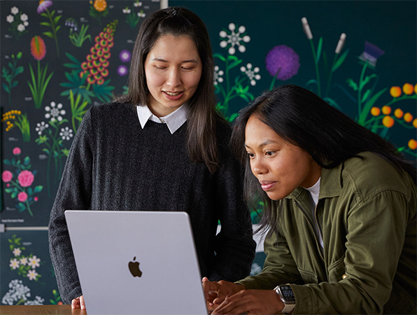 兩位 Apple 市場推廣團隊成員在其團隊設計的海報前，一起使用 MacBook 工作。
