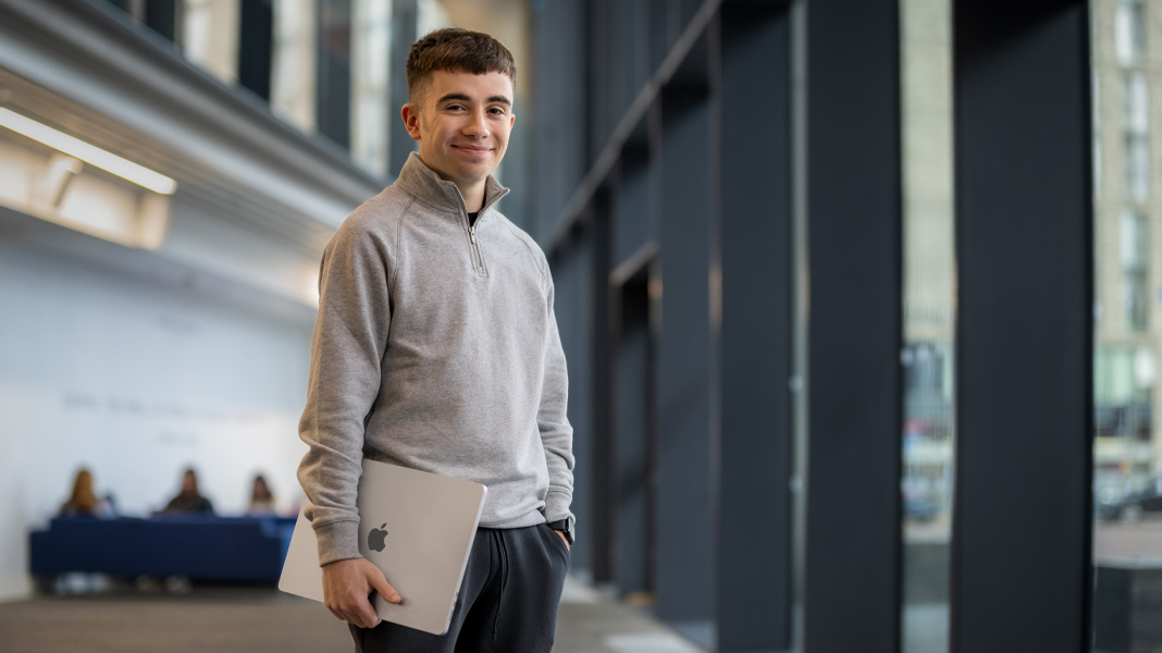En elev står i en moderne bygning, holder en bærbar MacBook, og smiler til kameraet