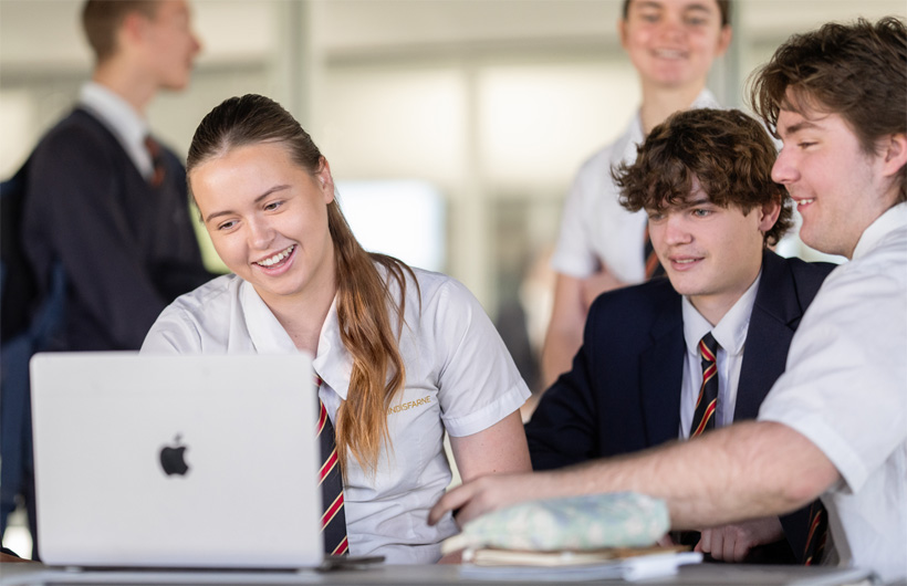 Um grupo de estudantes com uniformes escolares reúne-se à volta de um MacBook, a sorrir e a colaborar.