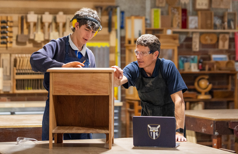 Um professor de marcenaria e um aluno analisam um pequeno armário de madeira. Existe um portátil escolar na bancada de trabalho à frente deles.
